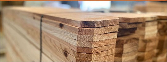 A close-up view of several stacked wooden planks with a smooth finish. The edges are neatly aligned, and the grain patterns are visible, indicating sturdy construction material in a workshop setting.