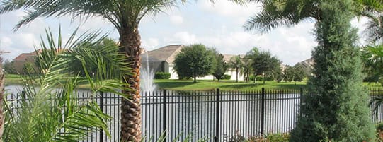 Pond surrounded by black fence, palm trees, and greenery. A fountain sprays water in the center. Houses with sloped roofs and trees are visible in the background under a partly cloudy sky.