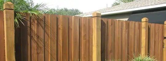 A wooden fence with vertical planks and decorative posts stands in front of a house with a shingled roof. Green plants slightly overhang the fence, and the sky is clear.