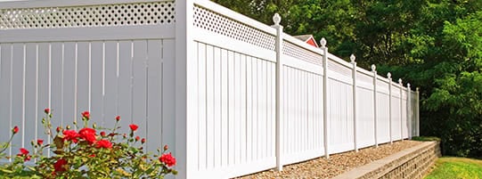 A long, white privacy fence with decorative lattice tops runs along a neatly landscaped area. Red flowers and green foliage are in the foreground, and the fence is built on a slight elevation with a stone border.