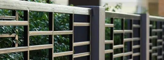 A gray metal fence with a rectangular pattern of bars runs alongside a black concrete wall. Green bushes are visible through the metal bars.