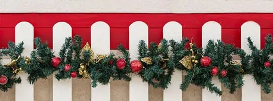 A festive garland with green pine branches, red ornaments, and gold decorations is draped over a white picket fence with a red background.