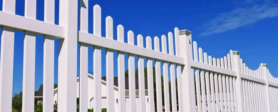 A white picket fence with vertical slats and decorative posts against a clear blue sky