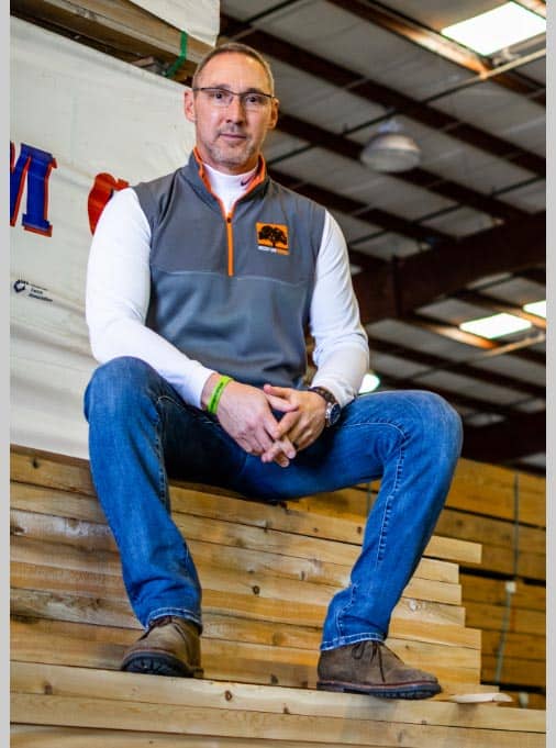 A man wearing glasses, a gray sleeveless vest, and jeans sits on a stack of wooden planks in an industrial warehouse. He is looking at the camera with a neutral expression. The background includes metal beams and a partially visible banner.