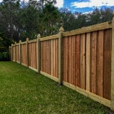 A tall wooden fence with vertical planks and capped posts runs across a grassy yard. Lush green trees are in the background under a partly cloudy blue sky.