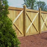A wooden privacy fence with diagonal cross braces stands on brown mulch, with a pine tree in the foreground and leafy trees in the background under a blue sky.