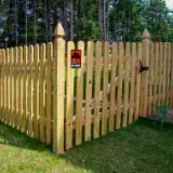 A wooden picket fence with a pointed top encloses a grassy area. A sign on the fence depicts a red dinosaur silhouette and the words "Clever Girl." Trees are visible in the background.