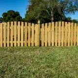 A wooden fence with tall planks sits on a grassy lawn under a clear blue sky. Trees are visible in the background.