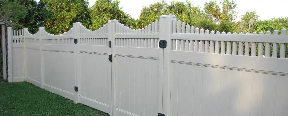White vinyl fence with alternating panel heights, featuring pointed tops and decorative curves, stands on grass with lush green trees in the background.