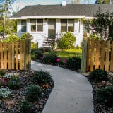 A quaint white house with a dark roof is seen beyond a wooden picket fence and a curved concrete pathway. The front yard is landscaped with various shrubs and colorful flowers, creating a welcoming garden entrance.
