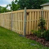A tall wooden fence runs along a grassy yard with freshly planted shrubs and flowers at its base. Trees and part of a house are visible in the background, under a partly cloudy sky.