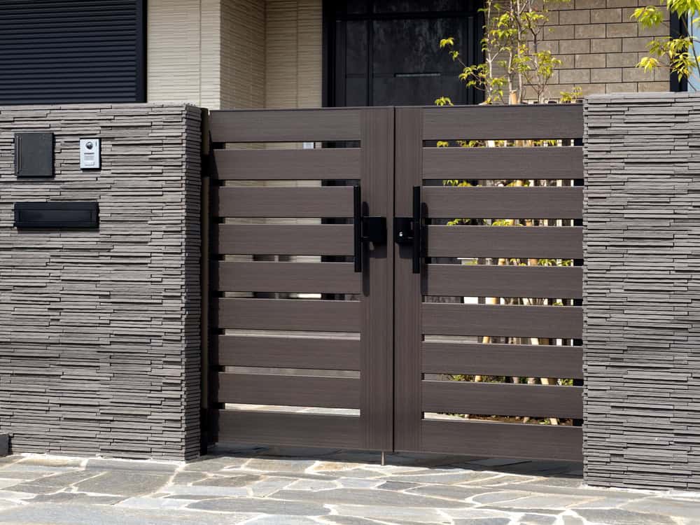 A modern wooden gate with horizontal slats set in a stone wall, featuring two black handles and an intercom on the left. The pathway in front is made of stone tiles. There are small green plants along the wall.