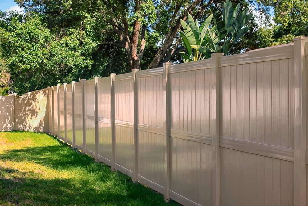 A long beige vinyl privacy fence runs alongside a grassy area, with lush green trees and large leaves visible in the background under a sunny sky.