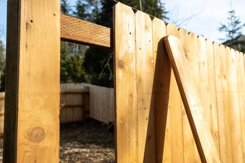 Close-up of a wooden fence under construction on a sunny day. The fence is missing some boards, revealing trees in the background. A narrow panel leans against the partially built structure.