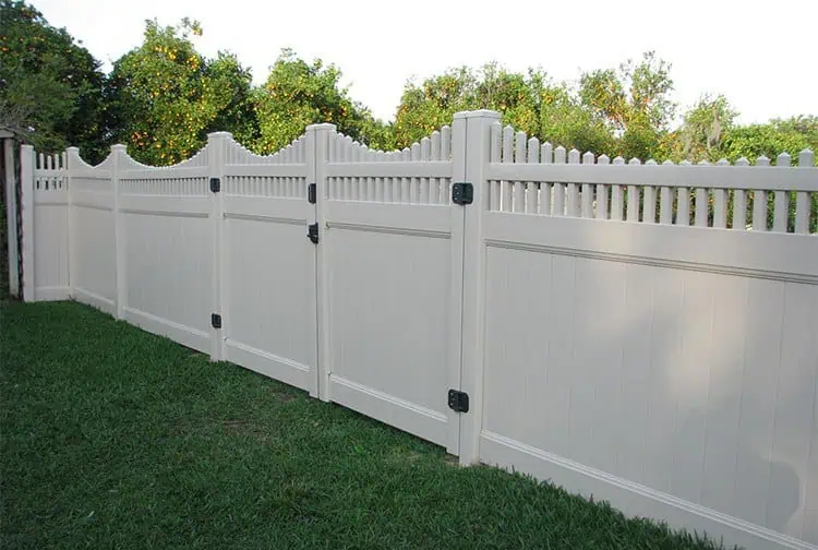 A white vinyl privacy fence with a decorative scalloped top and vertical slats borders a green grassy area. Trees and shrubs are visible in the background.