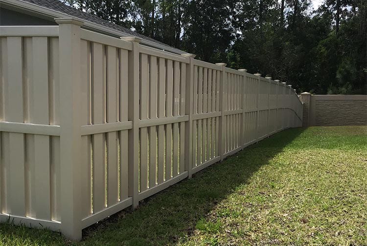 A long, beige vinyl privacy fence stretches alongside a manicured lawn. Tall trees form the backdrop, partially shading the fence and grass. The fence features vertical panels and is bordered by a corner towards the end.