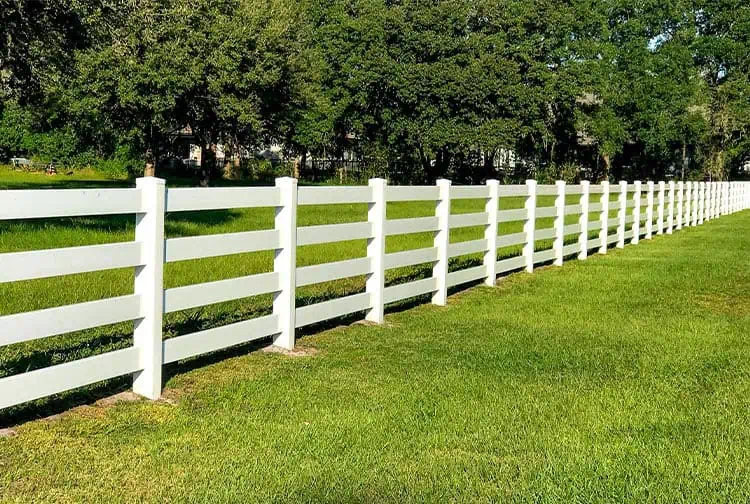 A long white fence stretches across a green grassy field, bordered by trees in the background under a clear blue sky.