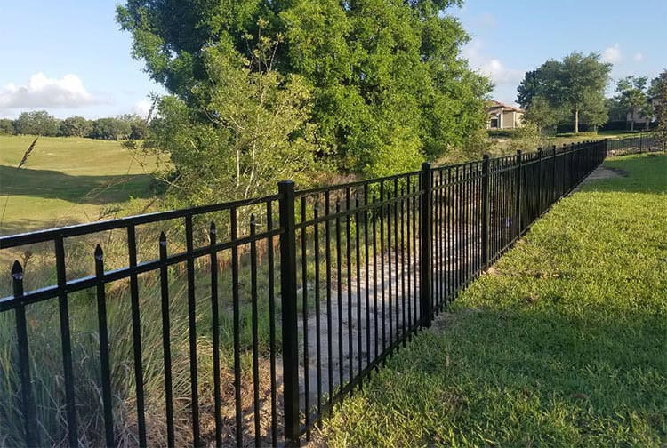 A long black metal fence stretches across a grassy landscape, with a path running alongside. Lush greenery and trees are in the background under a clear blue sky.