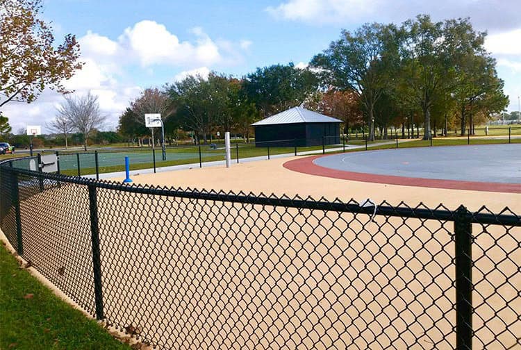 A fenced outdoor basketball court with a concrete surface, surrounded by trees and a clear sky. A small building with a metal roof is visible in the background.