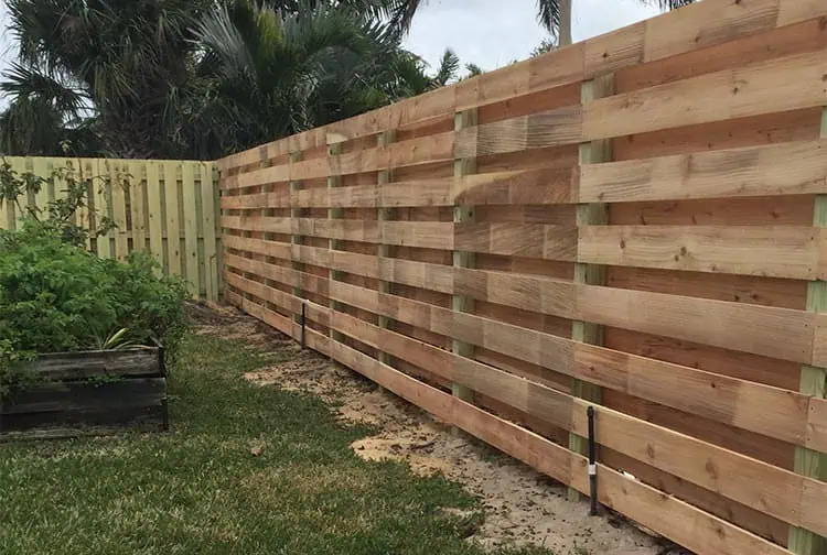 A backyard with a wooden horizontal slat fence and greenery in the background. The fence is newly built with natural wood tones, and there's grass and a raised, partially visible garden bed on the left.