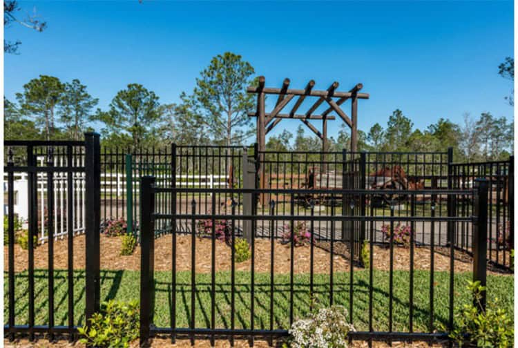 A black metal fence encloses a garden with mulch and small plants. In the background, there's a wooden arbor with a pathway leading through it. Tall trees and a clear blue sky are visible beyond the fence.