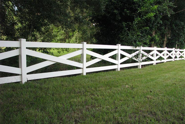 A long white wooden fence with a crisscross pattern is set against lush green grass and dense trees in the background, under a clear sky.