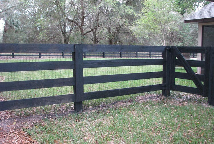 A black wooden fence with horizontal slats encloses a grassy yard. A gate on the right side is partially open. Trees and foliage are visible in the background.