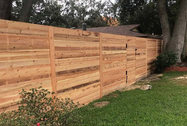 A tall wooden fence made of horizontal planks separates a grassy yard from a neighboring property. Trees and shrubbery are visible on both sides of the fence.