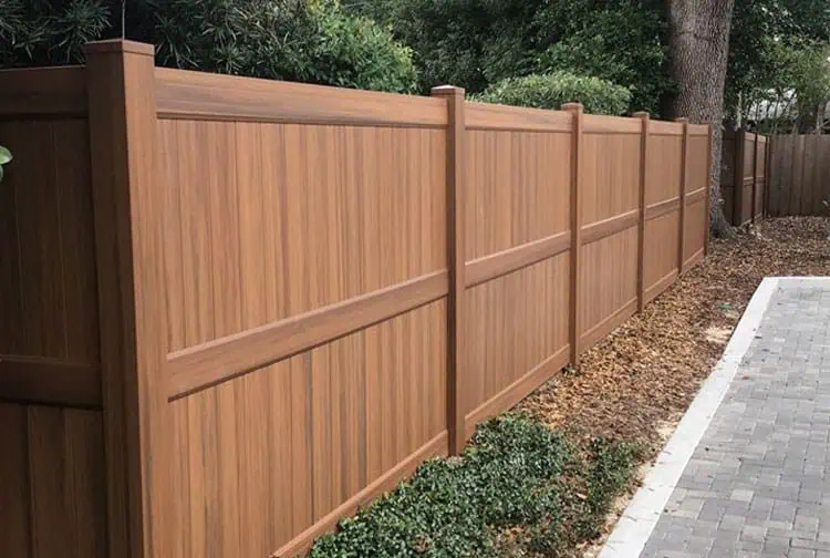 A wooden privacy fence lines the side of a yard, with tall, evenly spaced panels. The fence is surrounded by lush greenery and trees, and a paved pathway runs alongside the fence.