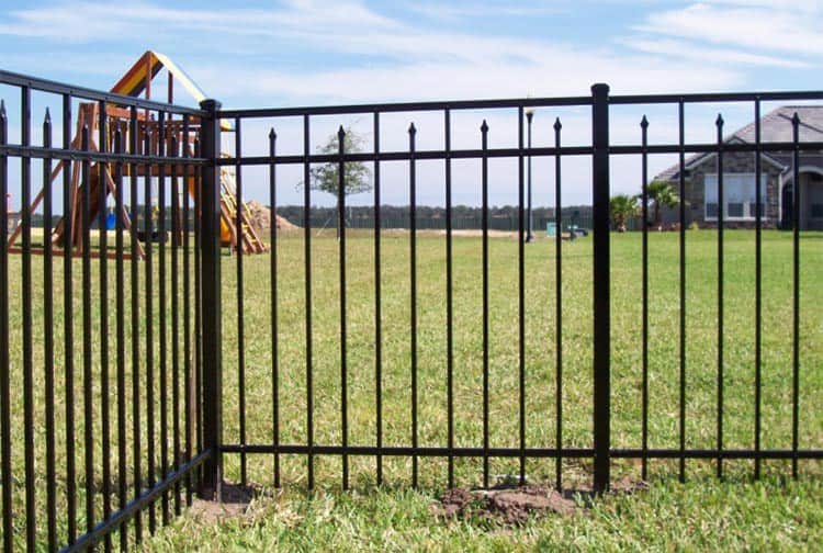 A grassy backyard with a black metal fence in the foreground. In the distance, a children's playset with a slide and swings is visible. A house is on the right, and the sky is partly cloudy.