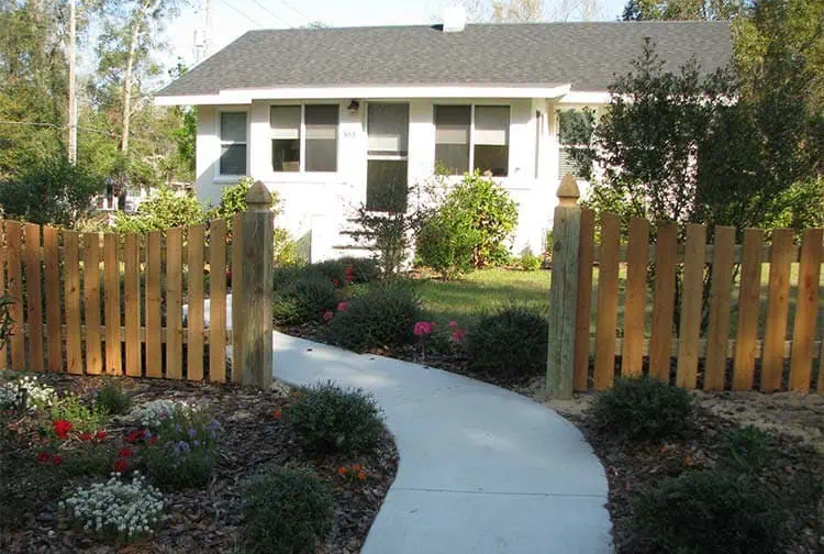 A small white house with a gray roof and a neatly cut lawn is behind a wooden picket fence. A curved concrete path lined with colorful flowers leads to the front door, surrounded by bushes and trees.