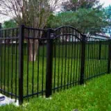 A black metal fence encloses a grassy area with trees in the background. The fence features vertical bars and an arched top section. The scene is outdoors, with greenery and a cloudy sky visible.