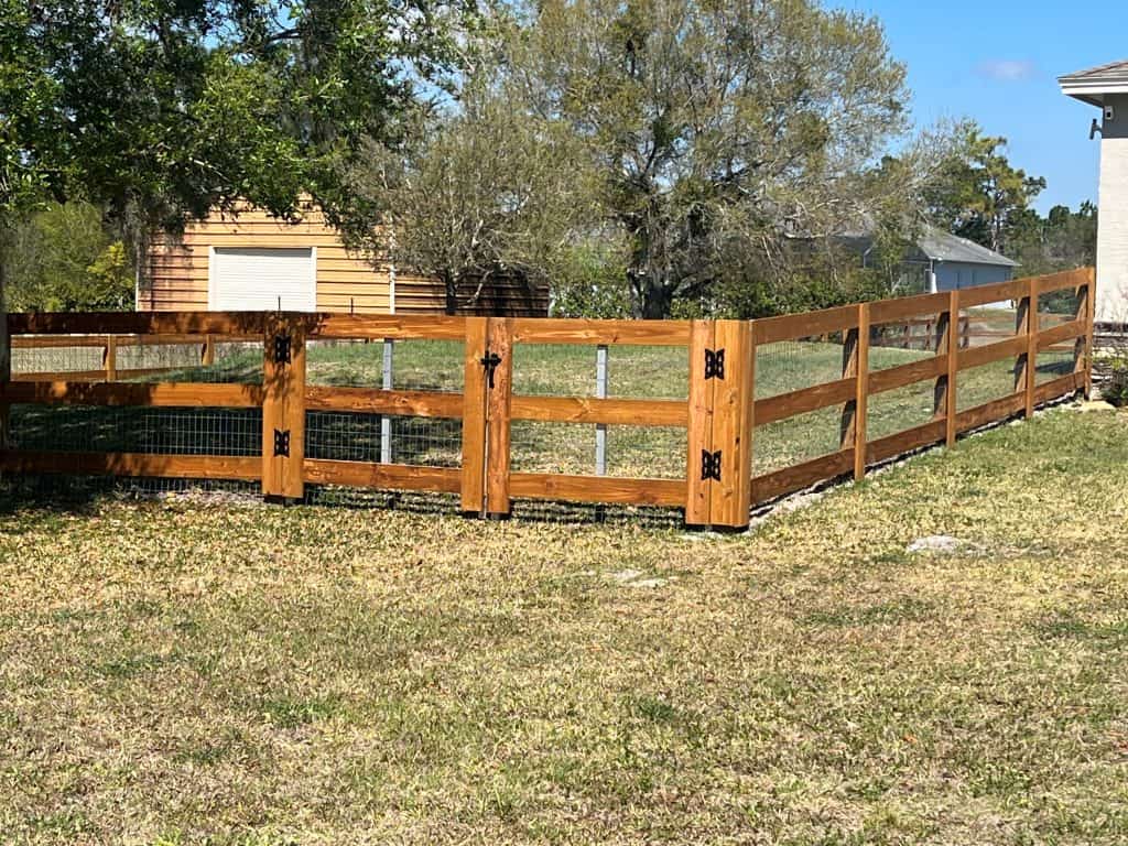 A wooden fence with metal mesh sections encloses a grassy yard. The fence features a gate with ornamental black hinges. Trees and a small building are in the background under a clear blue sky.