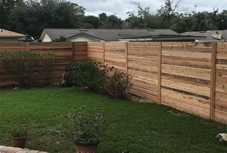 A backyard with a wooden privacy fence, green lawn, and several bushes and potted plants. Houses and trees are visible in the background under a cloudy sky.