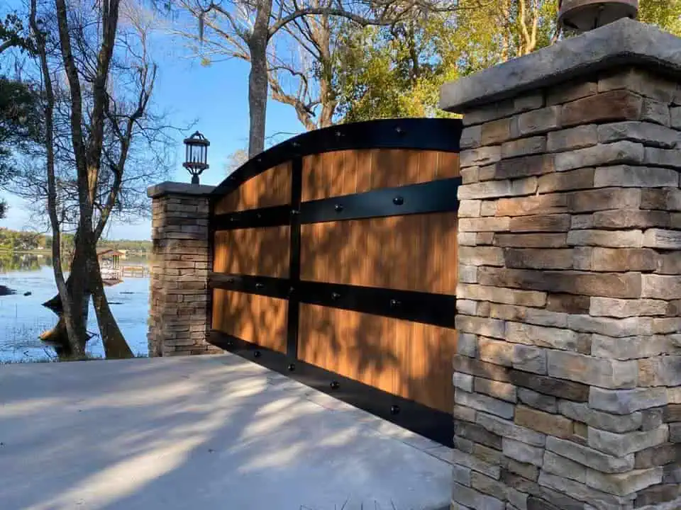 A stone pillar and wooden gate with metal accents stand beside a serene lake, surrounded by trees. A lantern adorns the top of the pillar, casting shadows on a sunny day.