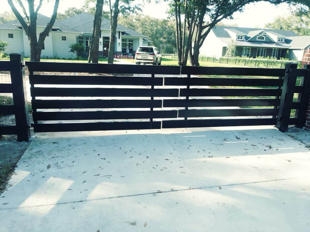 A modern, black wooden driveway gate stands closed at the end of a concrete driveway. In the background, there are two houses with trees surrounding them. Fallen leaves are scattered across the driveway.