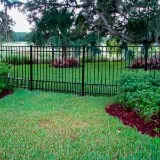 A well-maintained lawn with lush green grass is bordered by a black metal fence. The background features trees with dense foliage and ornamental plants, adding a touch of natural beauty to the scene.