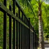 A close-up of a black metal fence with a pointed design, running alongside a path. In the background, there are lush green trees, including a tall palm tree, under bright sunlight. The image conveys a sense of serenity and nature.