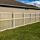 A beige wooden fence with vertical slats runs along the side of a grassy yard, adjacent to the beige wall of a house. The sky above is partly cloudy.