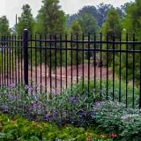 A black metal fence with vertical bars runs through a garden. In the foreground, colorful flowers and green plants are visible. Behind the fence, there are tall trees and an earthy landscape.