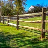 A wooden fence stretches across a grassy lawn, bordered by trees with some autumn foliage. A house with a white facade is visible in the background under a clear blue sky.