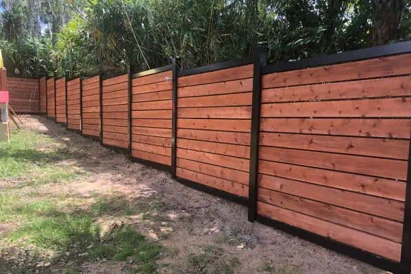 A long fence made of horizontal wooden planks with black metal posts stretches along a grassy area with tall plants in the background.