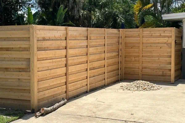 A tall wooden privacy fence encloses a backyard, with horizontal planks. A small pile of rocks is on a concrete slab in front of the fence, and a tree branch rests nearby. Lush greenery is visible over the top of the fence.