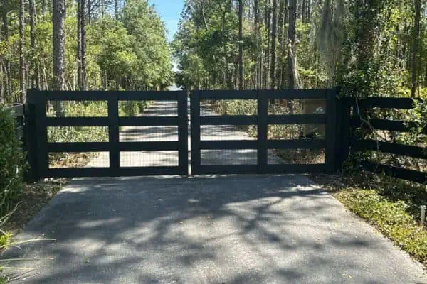 A wooden gate blocks a paved driveway surrounded by dense trees and greenery under a clear blue sky. Sunlight filters through the branches, casting shadows on the ground.