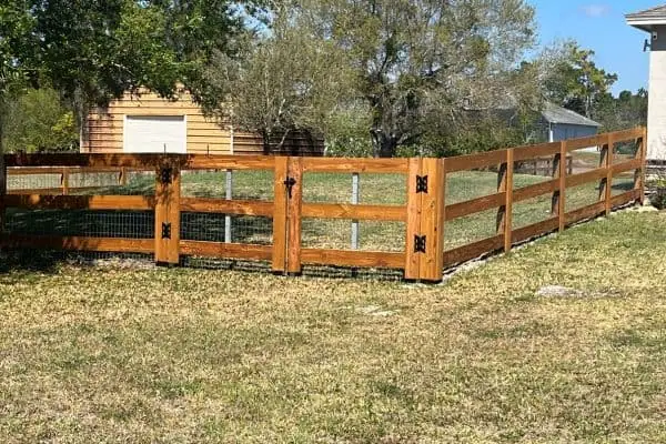 A wooden fence with metal mesh accents encloses a grassy area next to a house. The fence has decorative black hardware and is set against a backdrop of trees and a small wooden shed. The sky is clear and blue.