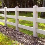 A beige wooden fence with horizontal slats and square posts runs parallel to a patch of mulch and grass. Trees with green foliage are visible in the background under dappled sunlight.