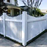 A white picket fence borders a suburban yard, with a two-story house in the background. The fence meets at a corner and runs alongside a concrete sidewalk. There are trees and a manicured lawn visible inside the enclosure.