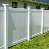 A white vinyl privacy fence with vertical panels and decorative posts runs alongside a house. The grass in front of the fence is green and well-maintained. The house's light-colored wall and windows are partially visible above the fence.