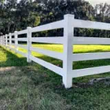 A long white wooden fence runs diagonally across a grassy field, with sunlight casting shadows. Tall trees with dense foliage are in the background under a clear sky.