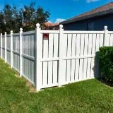 A white vinyl fence encloses a backyard with well-maintained green grass. The fence features vertical panels with decorative caps on the posts. A house is visible in the background, and a clear blue sky is overhead.